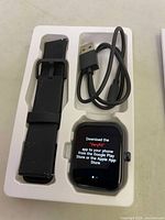 Smart watch device, black silicone strap, USB charging cable in white plastic tray packaging