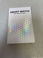 Photo of the front of white packaging box with holographic triangle pattern and text "SMART WATCH Your Sports Partner."