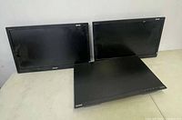 Three black computer monitors on a table; two standing vertically and one laying flat, showing front screen and bezels.