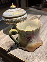 Medium white ceramic covered decorative bowl with multicolor floral designs and gold accents, with matching lid featuring a gold finial top; bowl has shaped rims and multicolor floral art with fading and wear visible indicating antique age.