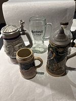 A group photo showing four drinkware items: two ceramic beer steins with pewter lids, one Avon ceramic mug, and one clear glass beer mug.