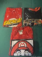 Three T-shirts folded and wrapped in clear plastic; red Bazinga! print, black Star Wars themed, and red Super Mario print shirts.