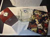 Brown wooden cigar humidor, clear square glass ashtray, and folded colorful Ferruche brand shirt with musical pattern on white surface.