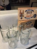 Four clear glass beer mugs and two beer tasting kit boxes with design and instructions, arranged on a white tablecloth.
