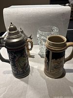 Two ceramic German beer steins with ornate pewter lids and one clear glass beer mug displayed on a dark surface.