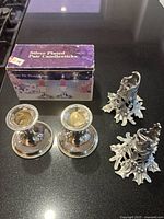 Photo showing two pairs of candlestick holders; one pair silver-plated with beaded edges and smaller in size, and one pair pewter with detailed holly leaf design. Original box visible in background.
