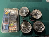 Four round fog lights with clear ribbed glass lenses and black metal housings arranged on table with remote starter kit in plastic packaging.