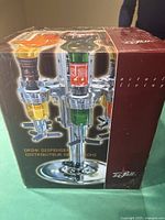Front packaging showing the assembled drink dispenser with three bottles inserted, chrome finish, base and dispensing taps visible.
