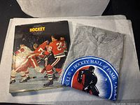 Photo of the Hockey Hall of Fame T-shirt folded next to a hockey book showing a vintage hockey scene on the cover.