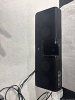 Front and side view of rectangular black Logitech iPod speaker and soundbar. Three speakers with protective mesh cover visible. Wired cord attached.
