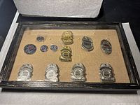 Full view of framed display with multiple metal guard and police badges, pins pinned to a beige fabric backing under glass.