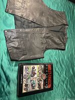 Photo showing the black leather motorcycle vest laid flat with the Harley-Davidson Chronicle book placed below it on a green fabric background.