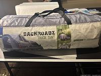 Photo of the tent in its storage bag, showing the model name Backroadz and displaying a large image of the tent attached to a pickup truck.