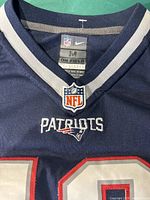 Close-up of jersey neckline showing NFL tag, size medium, 'On Field' label, and Patriots logo embroidered beneath collar.