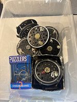 Plastic bin containing five vintage Pflueger fishing reels and one boxed metal chain puzzle toy.