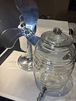 Metal retro fan with three polished blades and base next to a ribbed glass lemonade dispenser with lid and spigot.