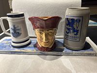 Photo showing three beer steins on a decorative blue and white serving tray. Left is a Royal Dalton stein with blue windmill design, center is a character jug stein with a red hat, right is a Löwenbräu München stoneware stein.