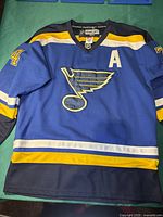 Front view of blue St. Louis Blues hockey jersey featuring number 74 on sleeves and the alternate captain 'A' patch. Shows vibrant team colors and logo.