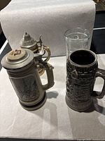 Four items including The Shipbuilder beer stein, two other ceramic beer steins with pewter lids, and a clear glass on a table.