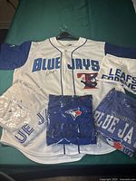Front of the signed Toronto Blue Jays white and blue jersey, with other jerseys and t-shirts folded around it on a green surface. The signed jersey has 'BLUE JAYS' text, the logo on the front and is size XL as stated.
