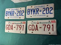 Four vintage metal Canadian license plates laid on a green fabric surface: two Ontario plates in white and blue and two New Brunswick plates in white and red with a date sticker '04/2006'.