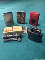 Five vintage metal lighters shown grouped on blue fabric background, including Dunhill lighter with original box, three flip-top lighters with designs, and one cylindrical lighter.