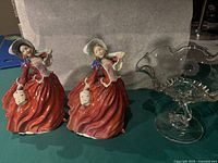 Two Royal Doulton figurines in red dresses with bonnets and one clear glass bowl with ruffled edge and pedestal base on a dark green surface
