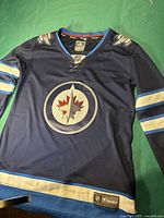 Front view of blue Winnipeg Jets hockey jersey with team logo and NHL 100th anniversary patches on shoulders.