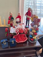 Four Byers Choice wooden and cloth caroler figures with a Santa decoration in the foreground, two small blue boxes on the table, and a tall glass vase filled with Christmas bulbs and fabric fruit ornaments beside them.