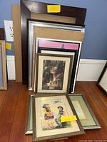Stack of approximately 8 framed wall art pieces leaning against a wall, showing various frame styles and sizes.
