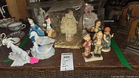 Collection of Hummel porcelain figurines on wooden surface including a dog, bird, and boxed figure in background