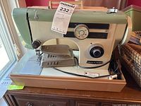 Vintage beige and green Oriccar sewing machine with control dials and case, placed on wooden surface next to window with natural light.