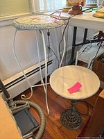 Two metal plant stands side by side. One has a floral mosaic stone top and white metal curved legs. The other has a dark metal base with a detachable marble-like top. Each has a pink sticker on top.