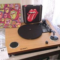 Front view of ION brand record player showing wood finished base, platter, tonearm, two built-in speakers, and a Rolling Stones-themed album cover in background.