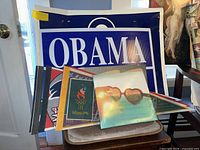 Photo of stacked pennants and signs with visible 'OBAMA' pennant and several sports-related pennants including an Olympic pennant. Items appear folded or stacked on a chair.