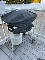 Weber propane grill front and side view with black Weber cover and propane tank underneath on a wooden deck with white fence.
