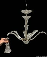 Chandelier shown hanging with six curved crystal arms, with one bell-shaped light cover held by hand, detachable from the arm.