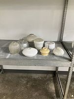 Eight art deco lamp glass shades and fixture glass displayed on a metal shelf, various shapes and finishes including ribbed, frosted and clear glass.