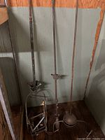 Photo of three vintage brass gas light fixtures against a wooden wall, including two tall straight pole fixtures and one incomplete harp fixture with square tubing.