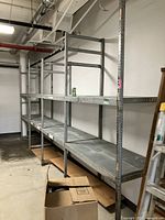 Four sections of heavy duty metal shelving, disassembled with three shelves each, placed against the wall in an industrial space.