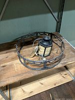 Photo shows black metal lantern placed inside a cast metal circular ring for lampshade on a wooden shelf.