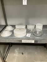 Shelf with eight lamp glass pieces: three milk glass flush mount dome shades, two shallow clear glass lamp shades, two clear glass lantern globes, and one milk glass tube.