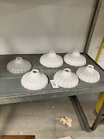 Six milk glass lamp shades displayed on a metal rack, showing varied sizes and shapes including larger and smaller ribbed and fluted designs.