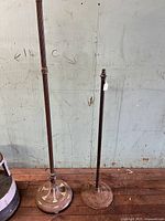 Two floor lamp bases with stems standing side by side against a light blue wall on concrete floor. Both metal, showing extensive rust and patina. One base wider and more decorative, the other thinner and simpler.