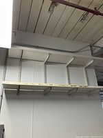 Long white wall shelf mounted on vertical rails with five heavy-duty white metal brackets on a wall in an industrial space.