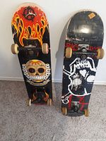 Two vintage complete skateboards standing upright, showing bottom deck graphics. The left skateboard has flames and sugar skull artwork, the right skateboard shows a skeleton holding a heart.