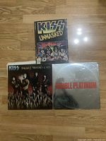 Top view of Kiss Unmasked book, Kiss Smashes Thrashes & Hits vinyl record, and Kiss Double Platinum vinyl record displayed on wooden floor.