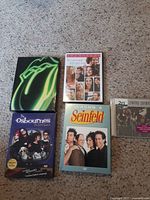 Top view of DVDs and CDs arranged on carpet including Seinfeld Season 5, The Osbournes Final Season, Let's Get Real with Lainie Diamond DVD, Lynyrd Skynyrd CD, and Rolling Stones box set.