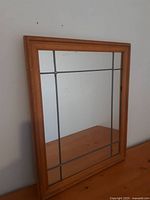 Full view of vintage rectangular mirror with wooden frame and grid pattern lines on the glass, leaning against a white wall on wooden surface.