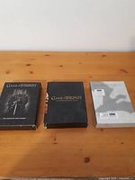 Three Game of Thrones DVD boxes displayed on a wooden surface: Season 1 with Iron Throne cover, Season 2 black box with gold lettering, and sealed Season 3 in plastic wrap.